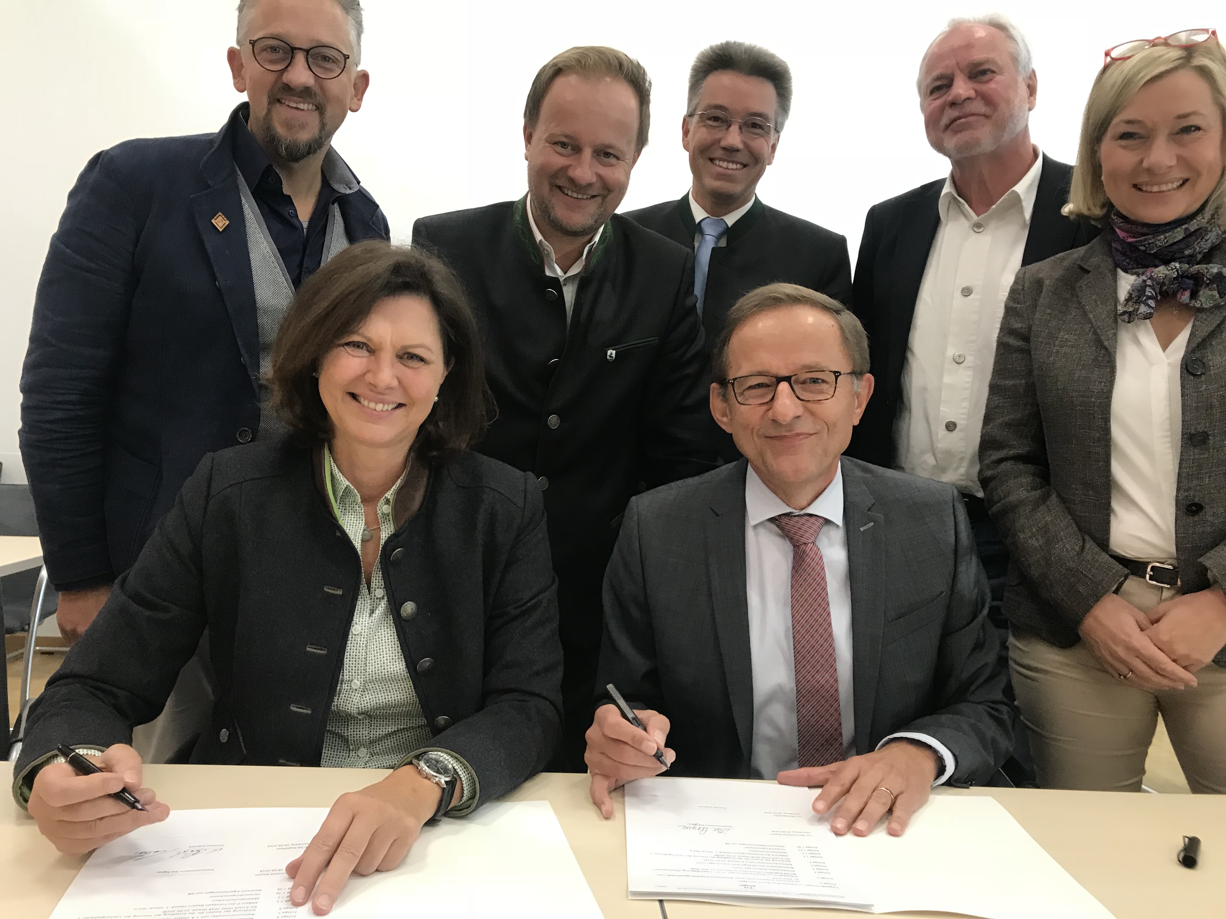 Ilse Aigner und Christian Kubasch sitzen an einem Tisch und unterzeichnen den Auftrag. Im Hintergrund stehen weitere Menschen.