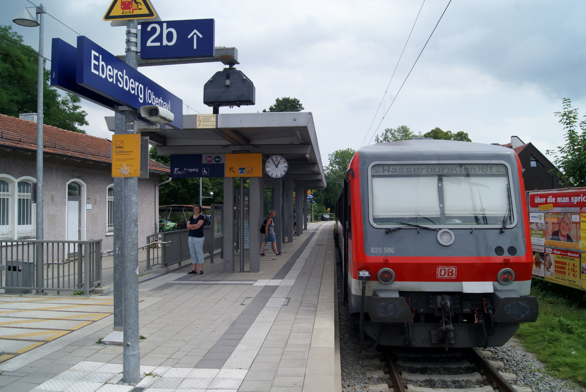 An der Station Ebersberg hält ein Dieseltriebwagen. Am Bahngleis stehen zwei Menschen und im Hintergrund ist das Bahnhofsgebäude.