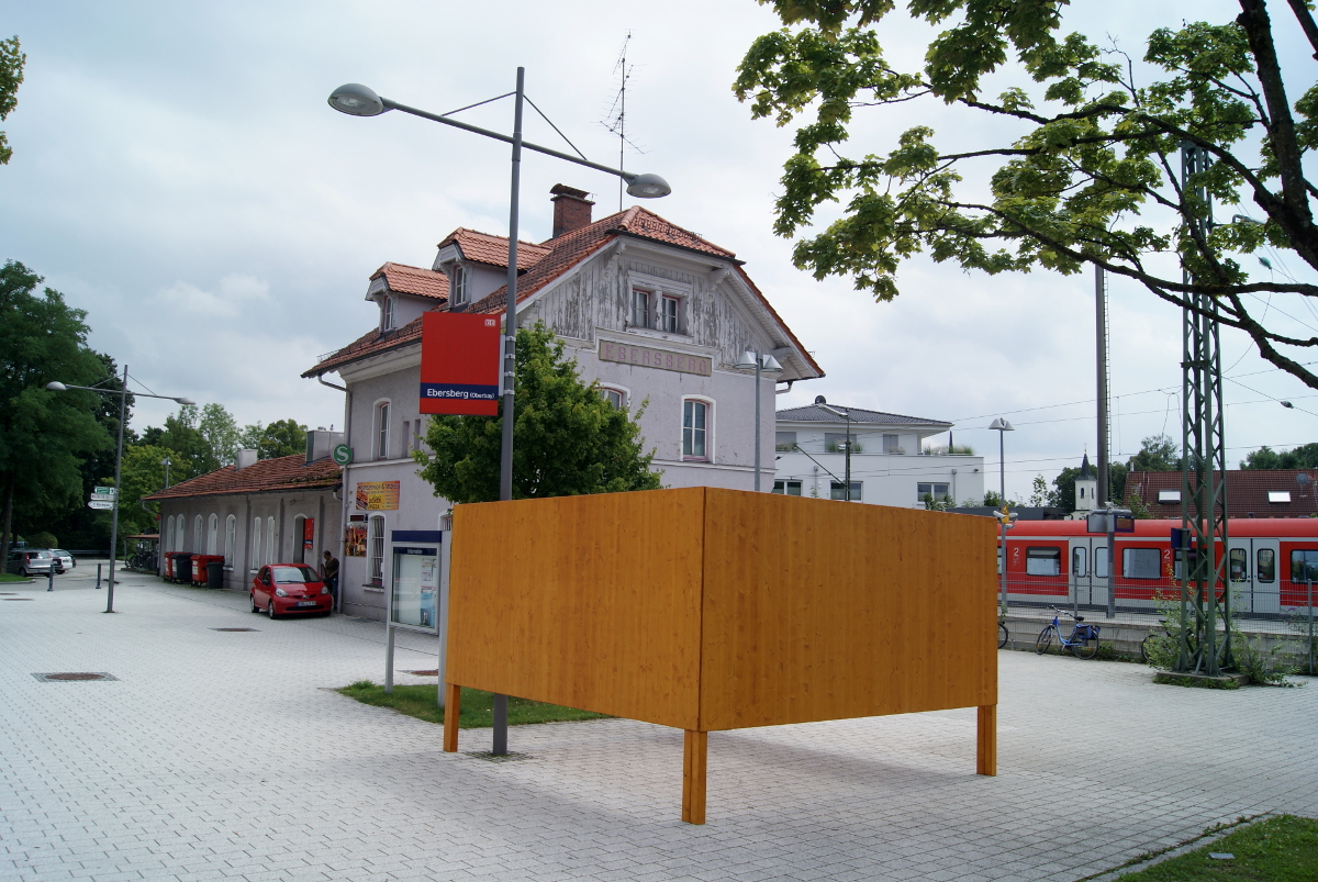 Vor dem Empfangsgebäude des Bahnhofs Ebersberg ist ein großer Platz. Vor dem Gebäude parken Autos und im Hintergrund sind Gleise.