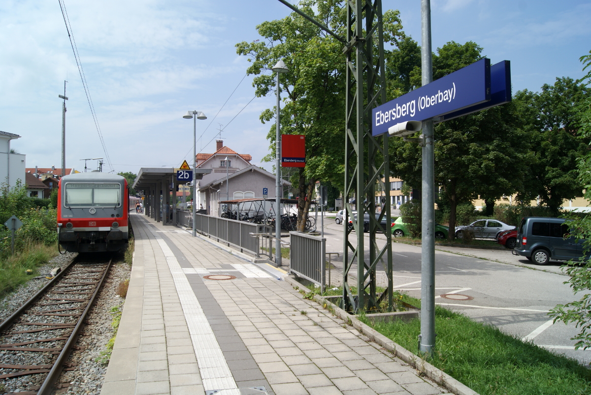 Ein Bahnsteig des Bahnhofs Ebersberg mit Bäumen und Autos im Hintergrund.