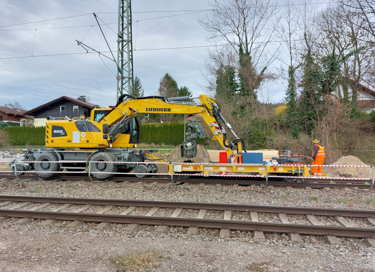 Auf der Baustelle arbeiten Fachkräfte in Schutzkleidung mit Hilfe eines Baggers weiter an der Baugrube neben den Schienen