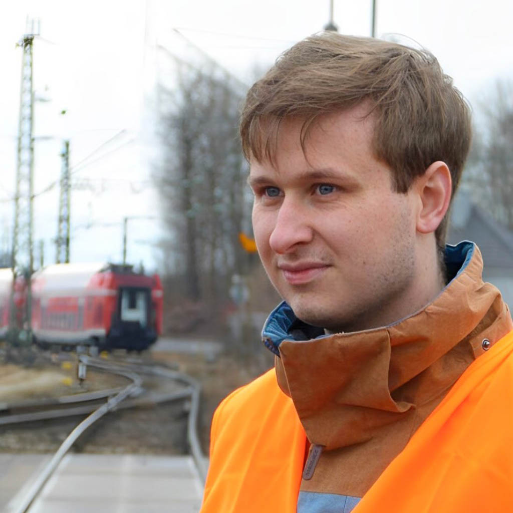 Lars Schindler an einer Eisenbahnstrecke, im Hintergrund eine rote Regionalbahn
