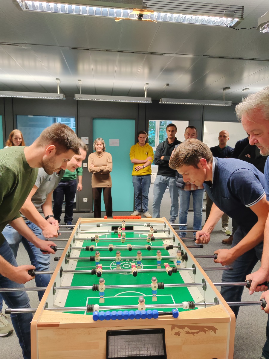 Zwei Zweier-Teams spielen gegeneinander Tischkicker. Im Hintergrund stehen weitere Mitarbeitende und schauen zu.