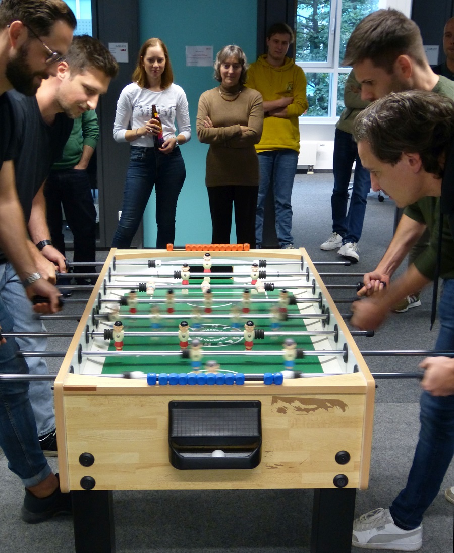 Zwei Teams beim Tischkicker spielen. Im Hintergrund stehen Menschen, die zuschauen.