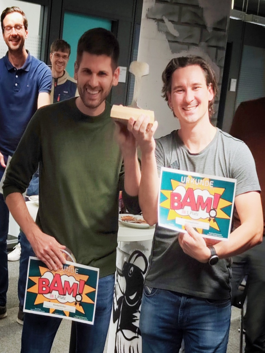 Foto von den zwei Gewinnern des Tischkicker-Turniers. Sie halten den Pokal, sowie jeweils eine BAM-Gewinner-Urkunde.