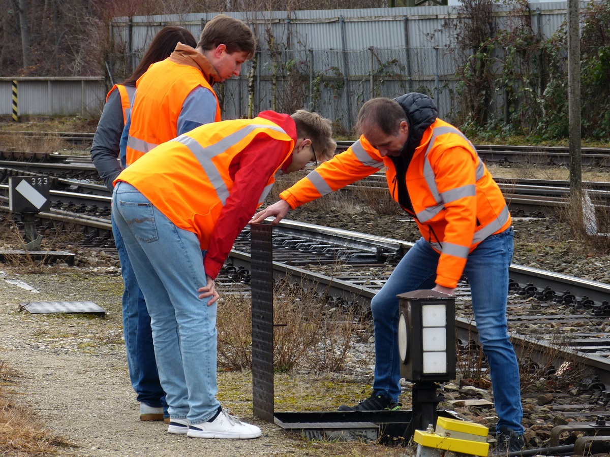 Ein Mensch in Schutzkleidung klappt eine Luke im Boden auf, während zwei andere Menschen ihm dabei zusehen.