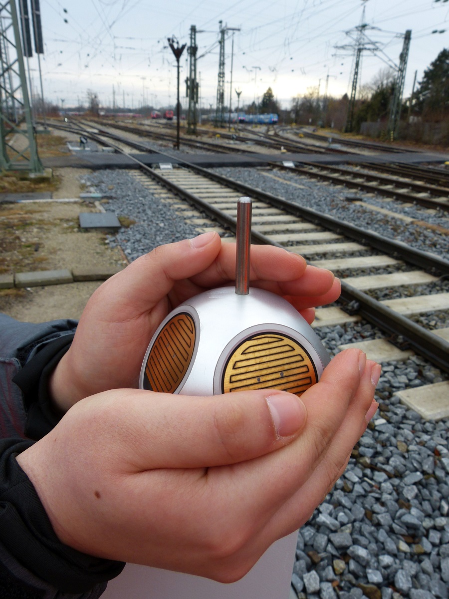 Zwei Hände halten ein kleines, viereckiges technisches Gerät, welches an den Gleisen angebracht werden soll.