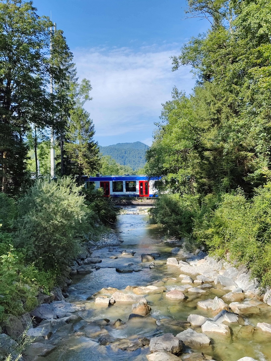 Landschaft mit Fluss und Bäumen, im Hintergrund fährt ein Regionalzug über eine Brücke.