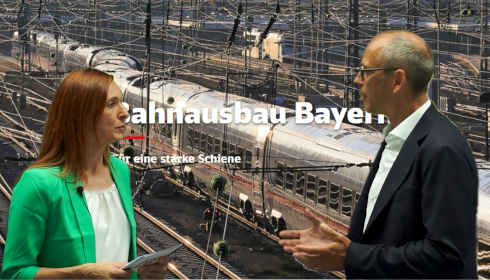 Zwei Mitarbeitende der Deutschen Bahn stehen vor einer Schienen-Grafik und unterhalten sich.